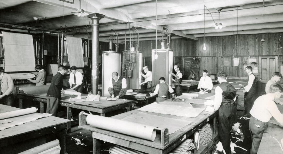 A black and white photo of men working on large tables in a factory or workshop. They appear to be handling large sheets of material, possibly for large format printing. The setting has industrial equipment and tools.