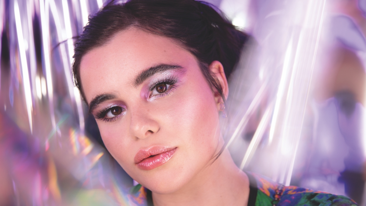 A person with glossy lips, shimmering eye makeup, and wearing colorful attire is looking directly at the camera in a well-lit environment with blurred background effects reminiscent of a high-quality 3D print.