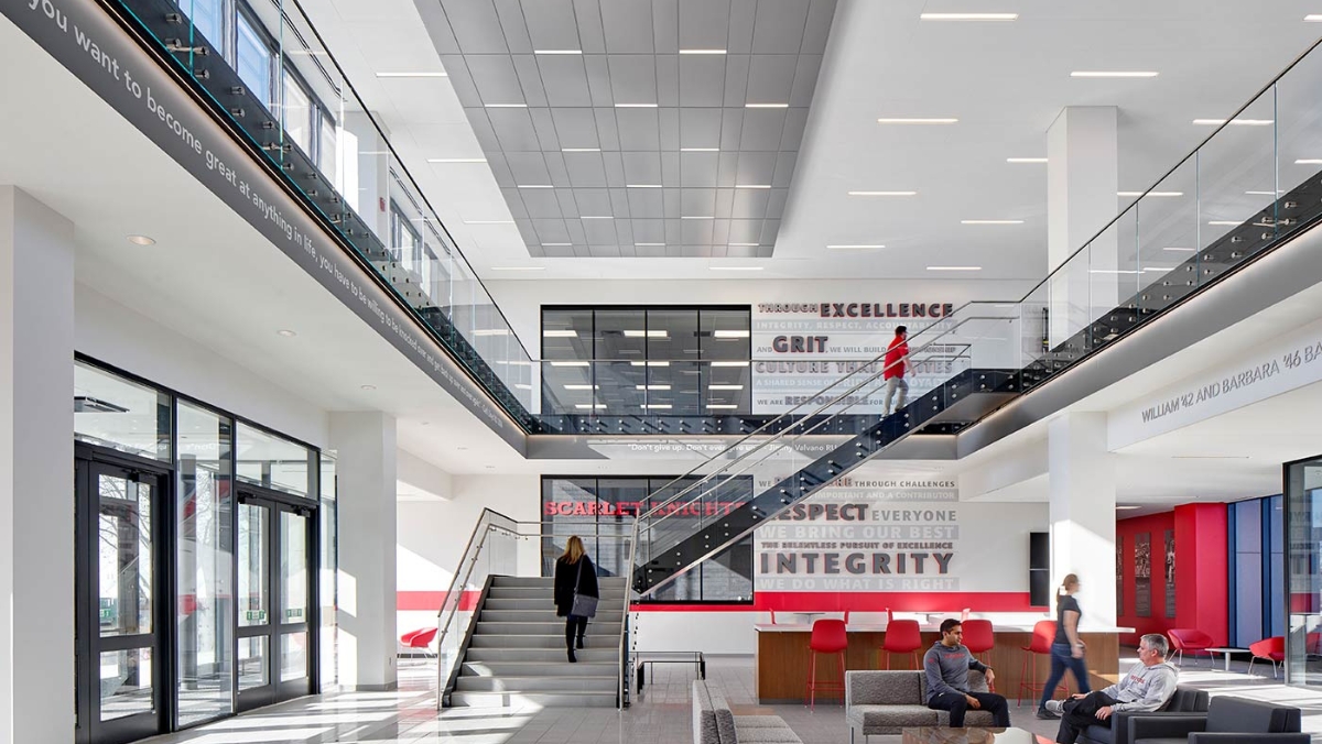 A modern, multi-story lobby with large windows and stairs leading to the second floor. Several people are walking and sitting in the space. Motivational words, likely created using large format printing, are displayed on the walls.