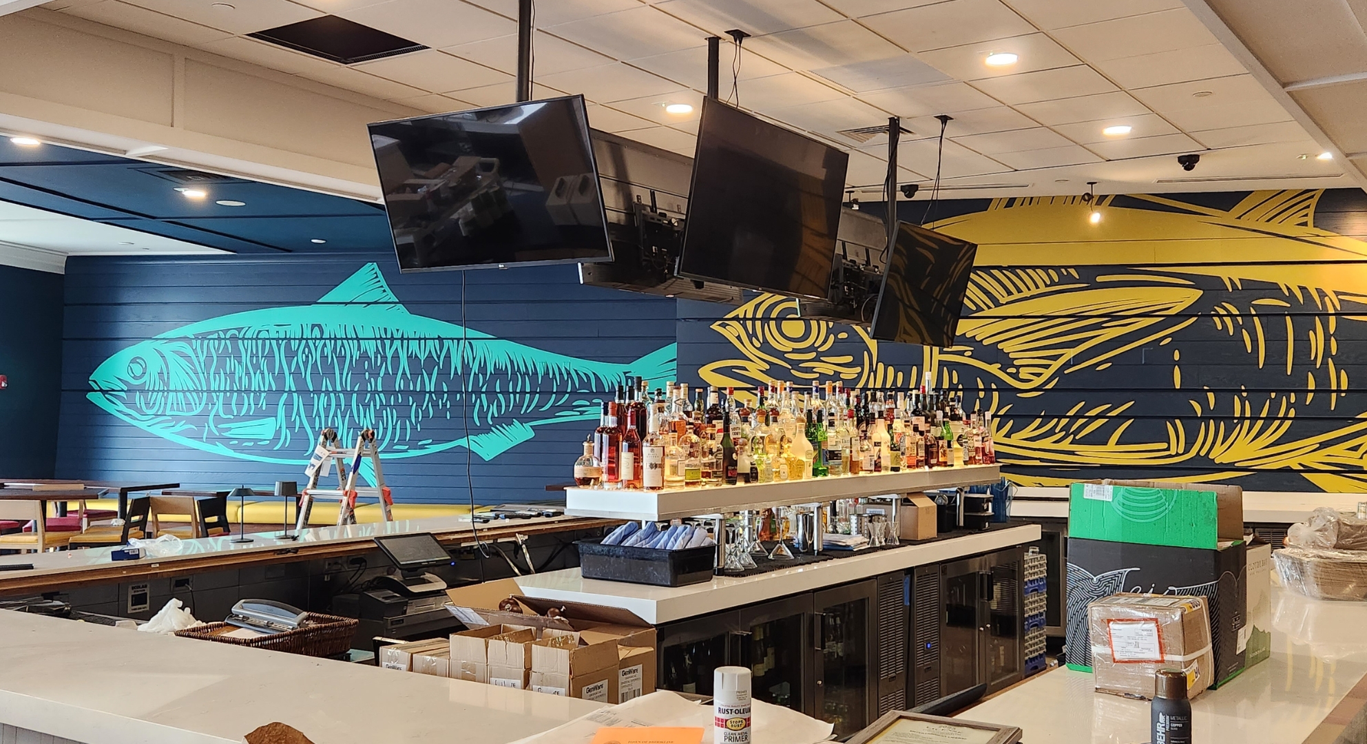 Interior of a bar with a counter full of various bottles, two overhead screens, and murals of fish in blue and yellow on the back wall, showcasing the elegance possible with large format printing.