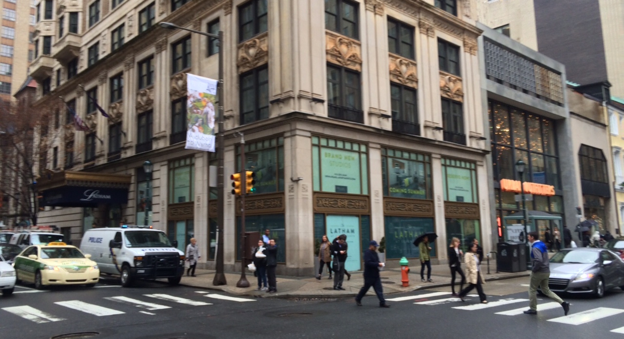 Pedestrians crossing a street corner in front of a multi-story building in a city. A police vehicle and other cars are parked near the intersection, possibly near a shop offering 3D printing services. The weather appears to be overcast.