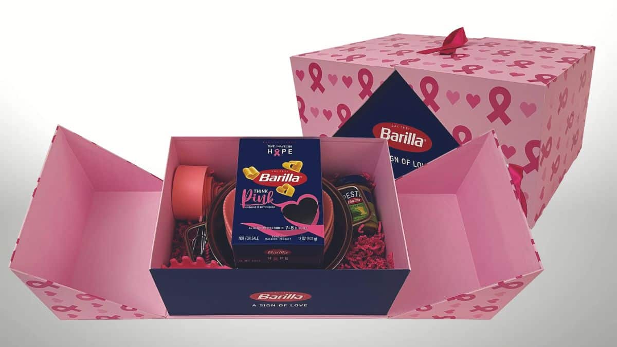 Breast cancer awareness gift box with pink ribbon decorations containing a pack of Barilla pasta, olive oil, tomato sauce, and pink kitchen utensils. The customized packaging incorporates large format printing to highlight the cause.