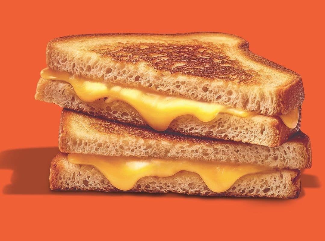 Two halves of a grilled cheese sandwich are stacked, showing melted cheese between toasted bread slices, against an orange background.