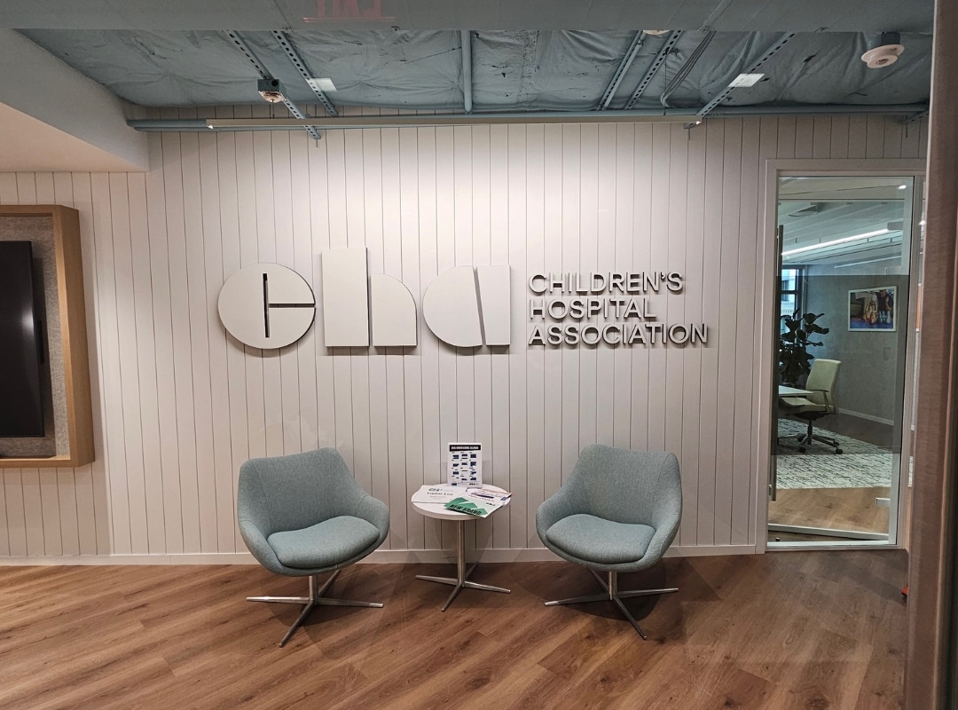 Two light blue chairs and a small table sit in front of a wall with the Children's Hospital Association logo and name in a modern office setting.