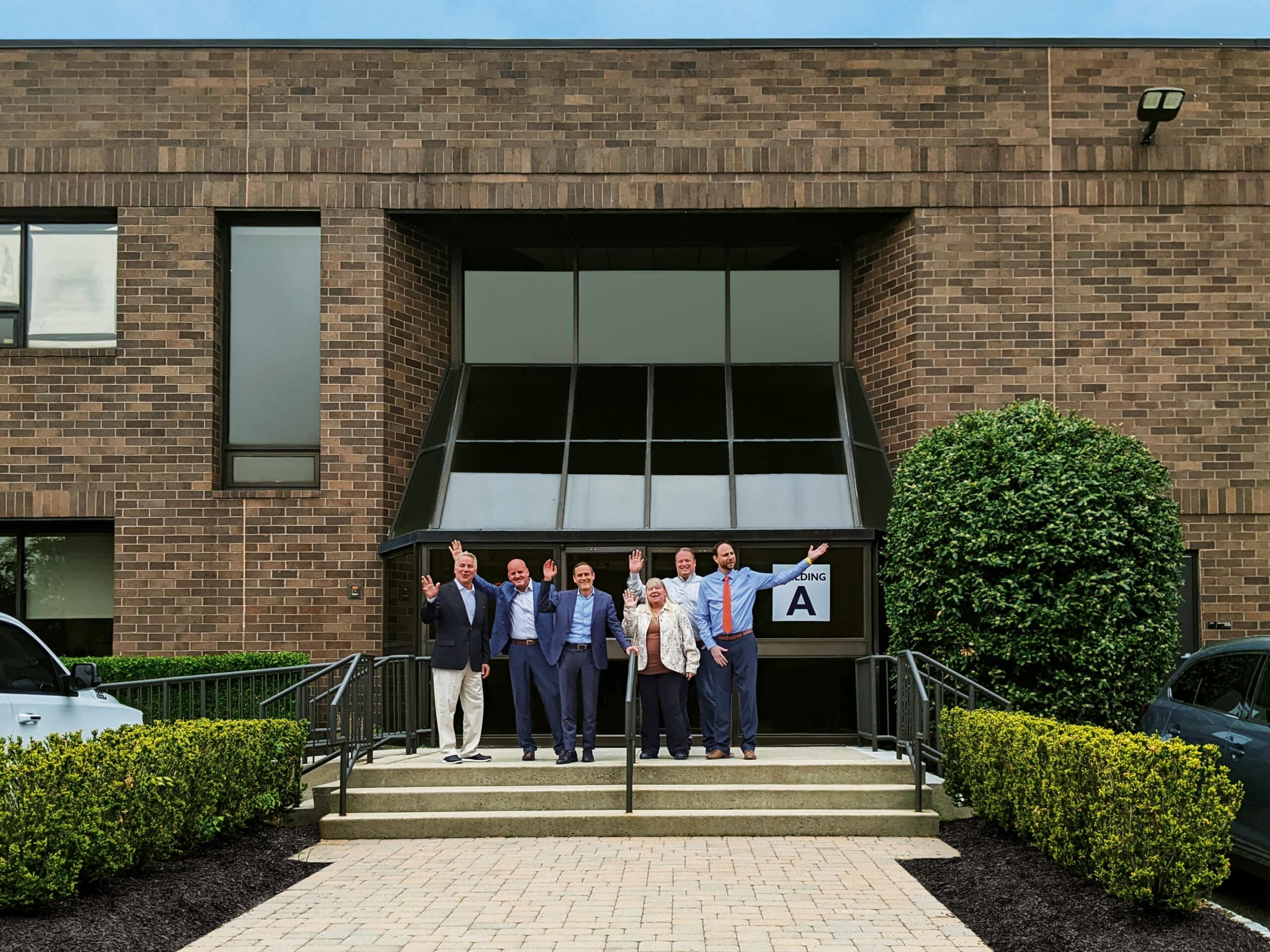 Seven people stand on the stairs outside a brick building with large windows, posing cheerfully, as if celebrating the latest achievement in their 3D printing services venture.