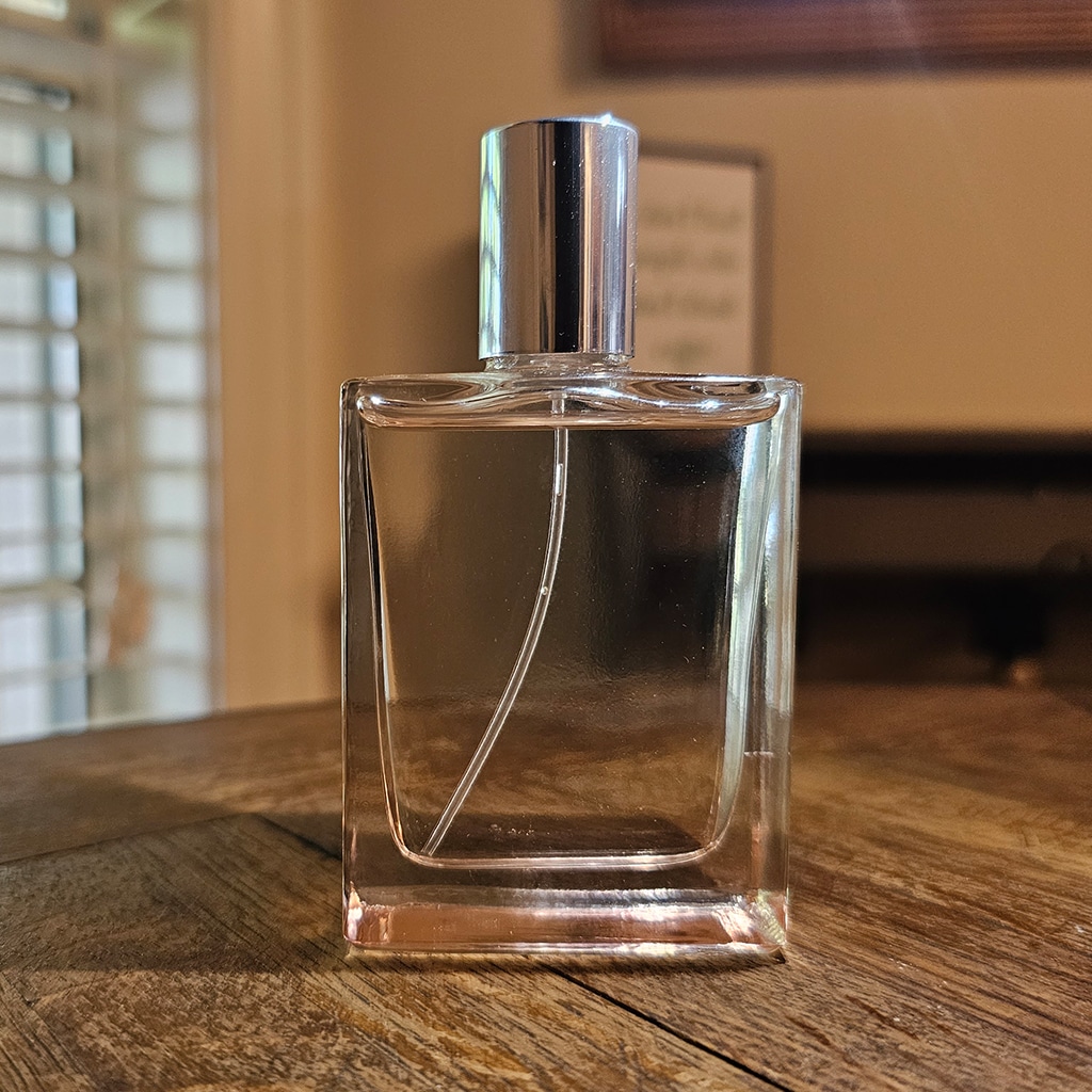 A clear glass perfume bottle with a silver cap sits on a wooden table.