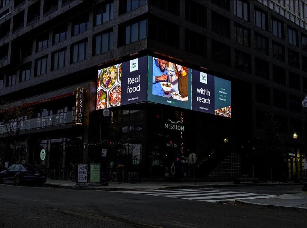 Large LED billboard on a building corner displays an ad for a food delivery service with images of meals and text "Real food within reach." Street and restaurant below are dimly lit.