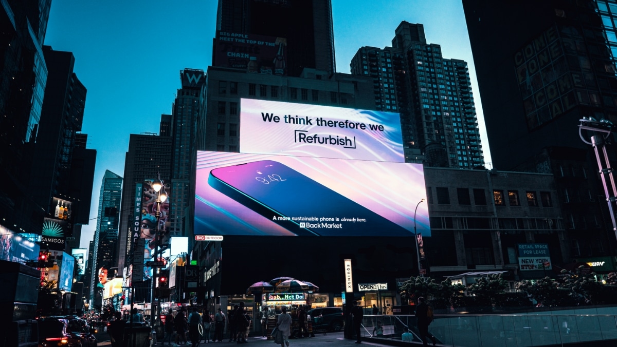 OOH Layouts Large electronic billboard in a city displays an advertisement for a refurbished phone with the slogan "We think therefore we Refurbish." Buildings and lights are visible in the background.