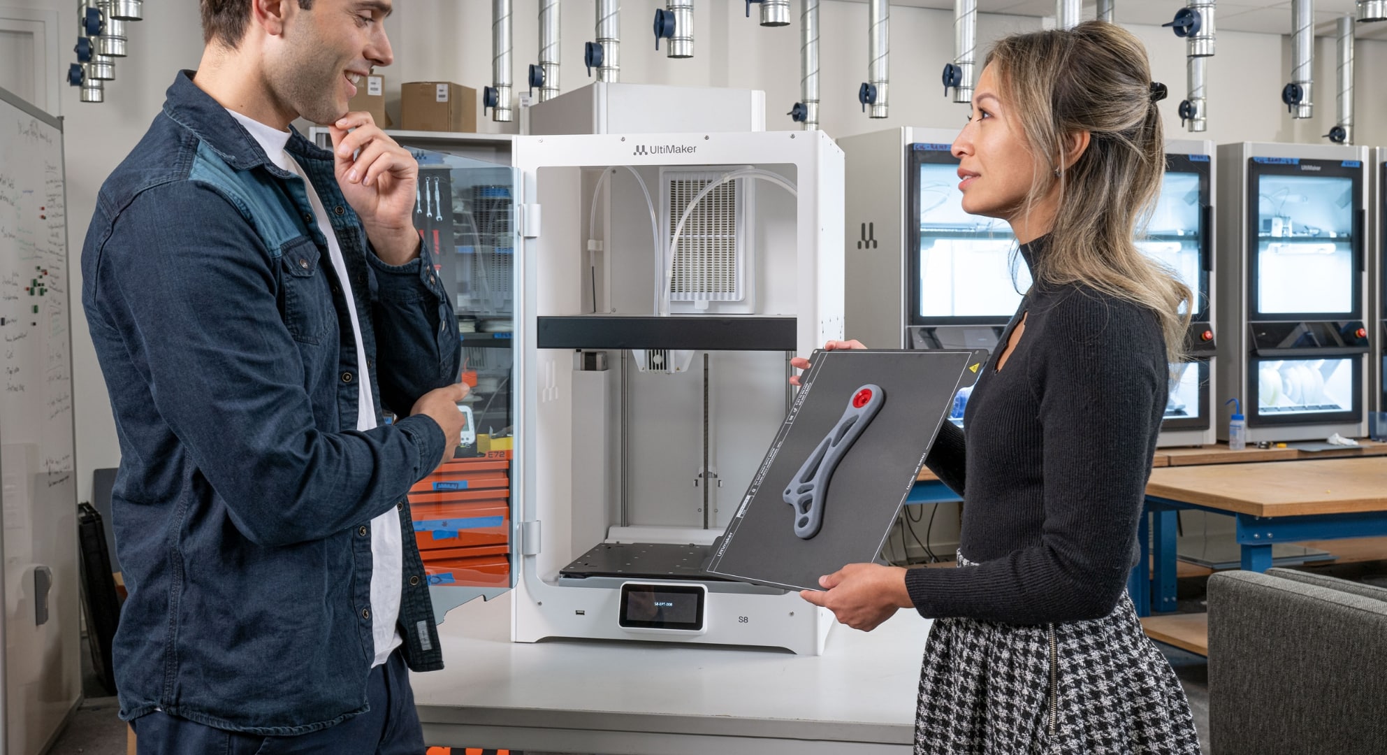 Two people stand in a lab discussing a 3D-printed part; the woman holds a tray with the part in front of a 3D printer.