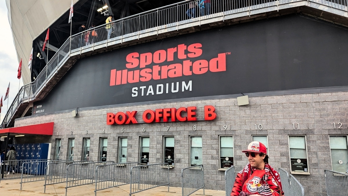 Red Bulls Large Format A person in sports fan attire stands outside the Sports Illustrated Stadium near Box Office B with numbered ticket windows in the background.