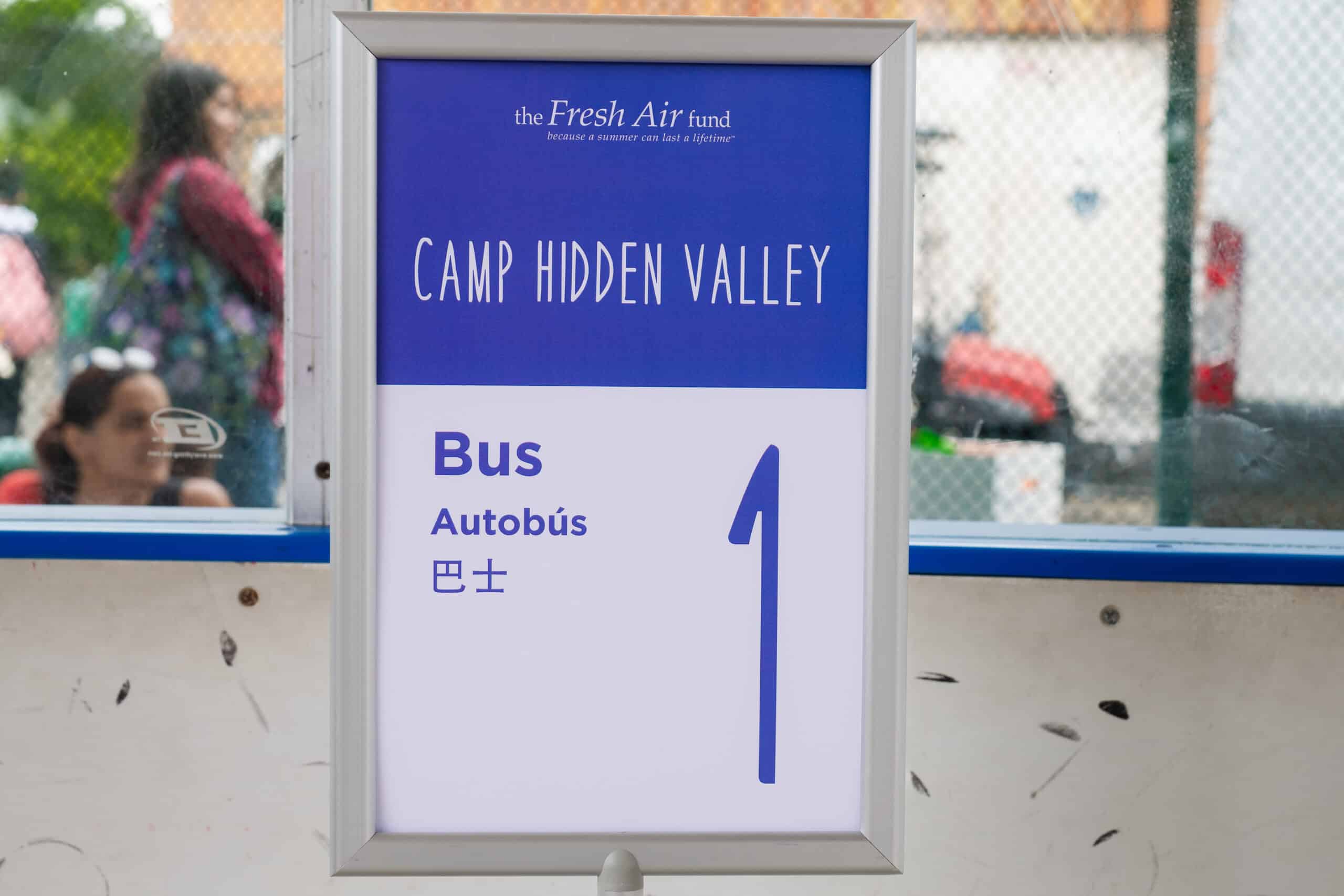 06.27.25.PC-TiyeRose_RiverbankDepartures_DSC05050 A sign reads "Camp Hidden Valley Bus 1" in English, Spanish, and Chinese, with "The Fresh Air Fund" written at the top. People and a fence are visible in the background.