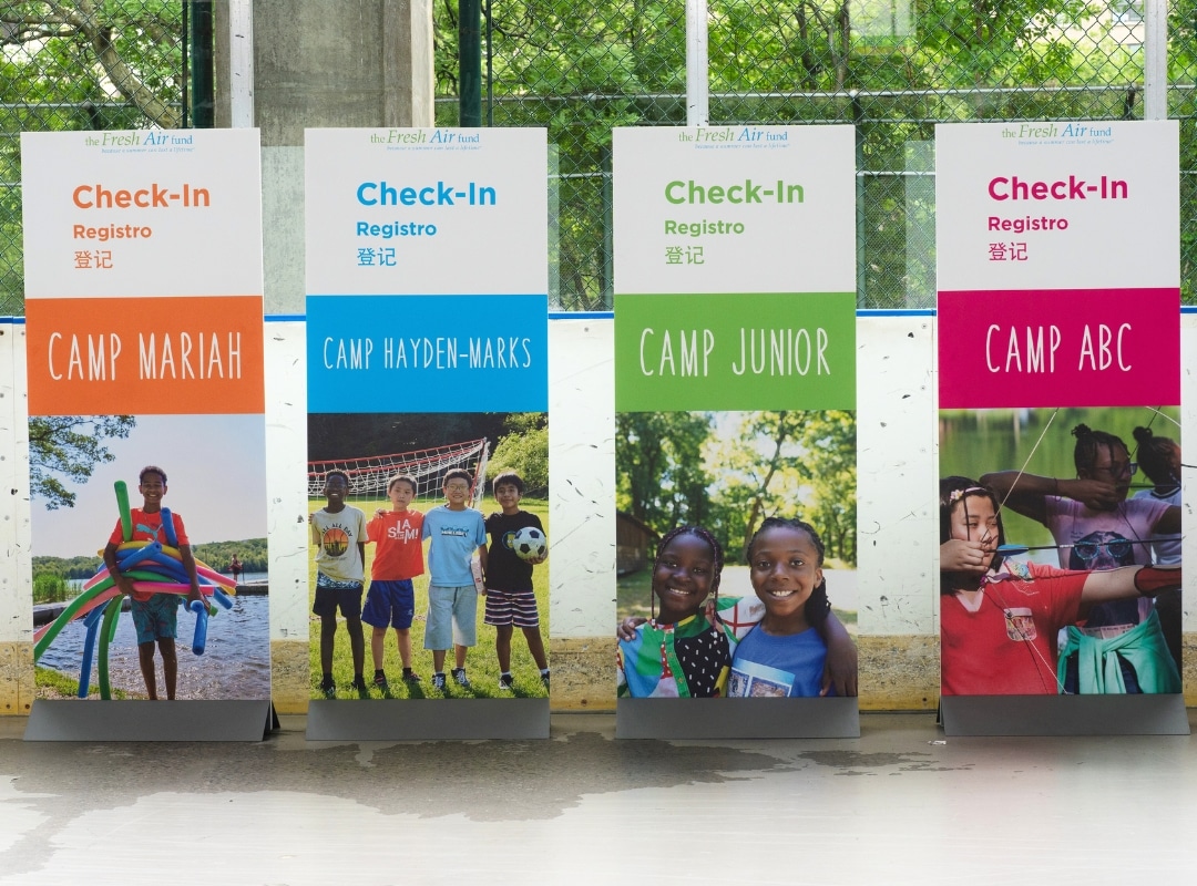 Four colorful camp check-in signs labeled Camp Mariah, Camp Hayden-Marks, Camp Junior, and Camp ABC, each with photos of children and bilingual English/Spanish text.