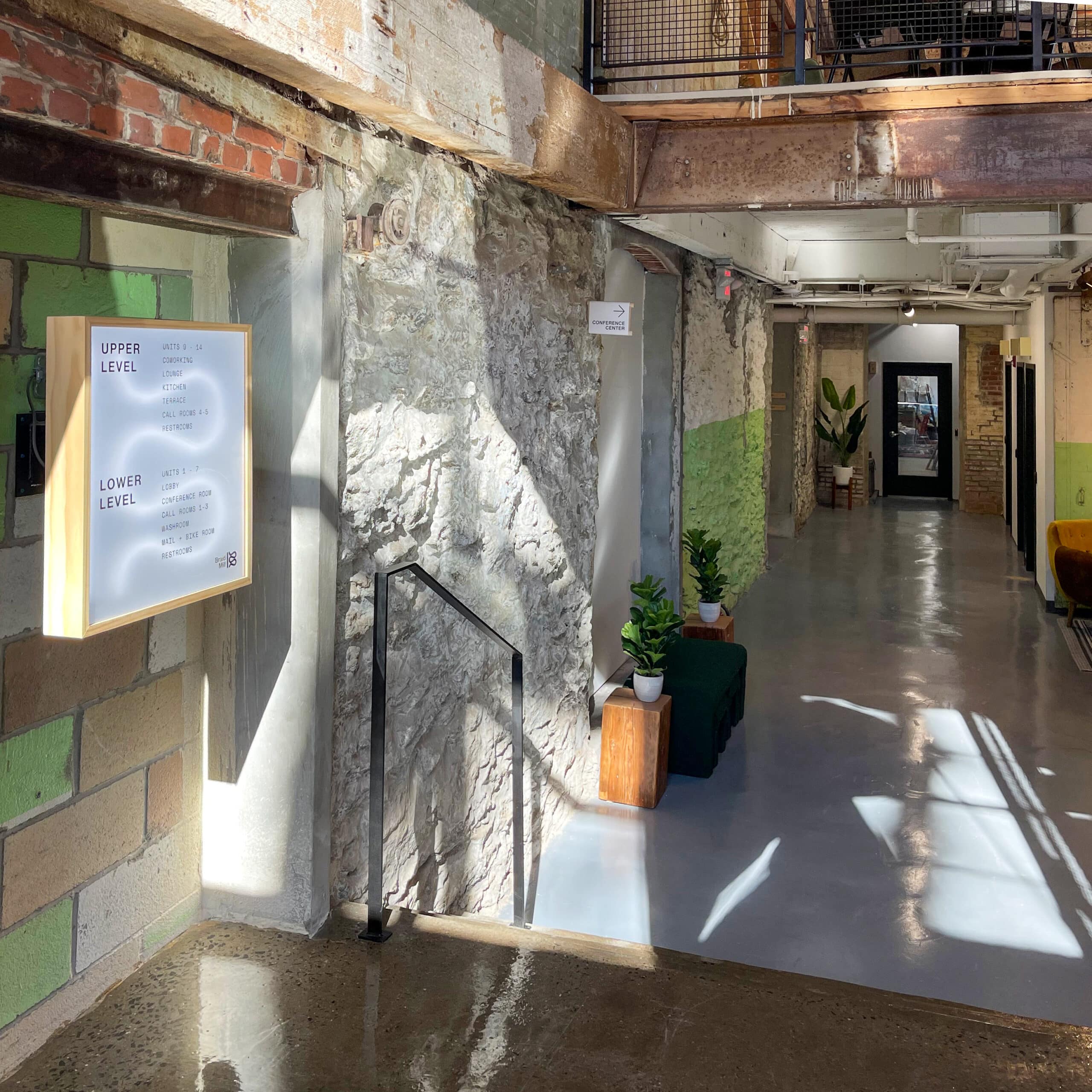 A well-lit corridor with a stone wall, a directory sign, stair railing, potted plants, and a glass door at the far end. Sunlight streams in, casting shadows on the floor.