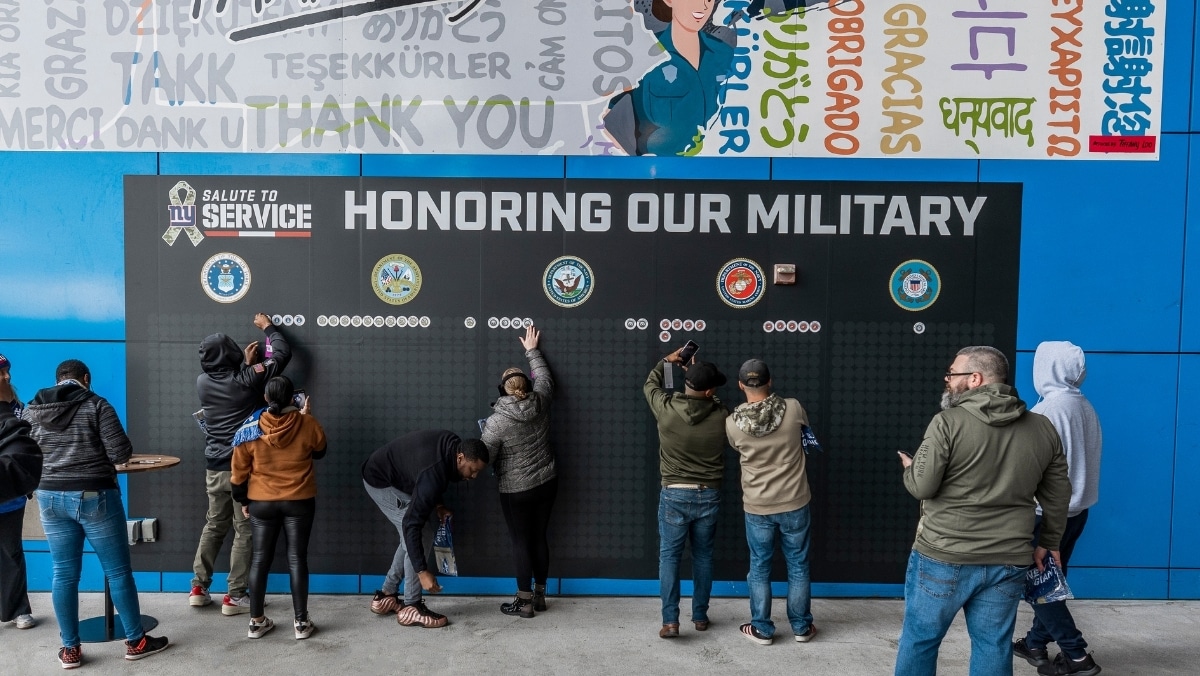 120 People place white hockey pucks on a display wall labeled "Honoring Our Military," under military branch logos, at an event with a mural saying "Thank you" in multiple languages.