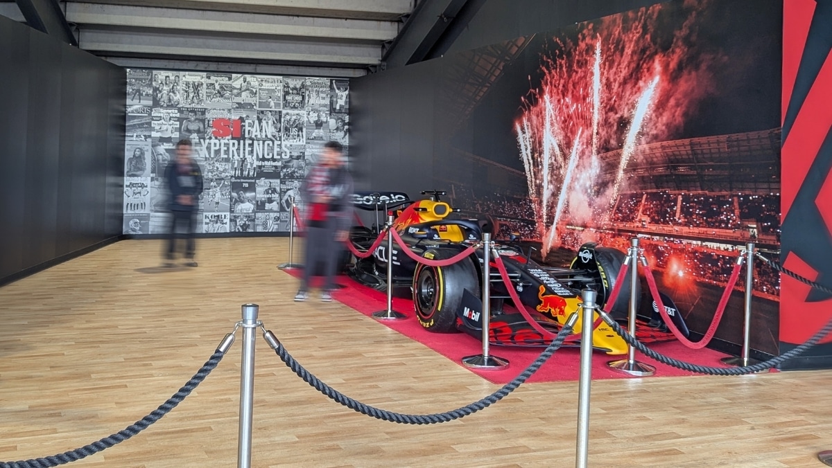 121 A Red Bull Formula 1 car is displayed on a red carpet, roped off, with a backdrop showing fireworks and stadium lights; two blurred people stand nearby.