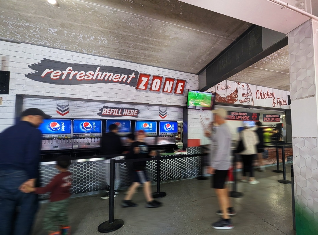 People stand in line at a refreshment zone with Pepsi refill machines, next to a food counter offering chicken and fries; a TV displays a sports game above.