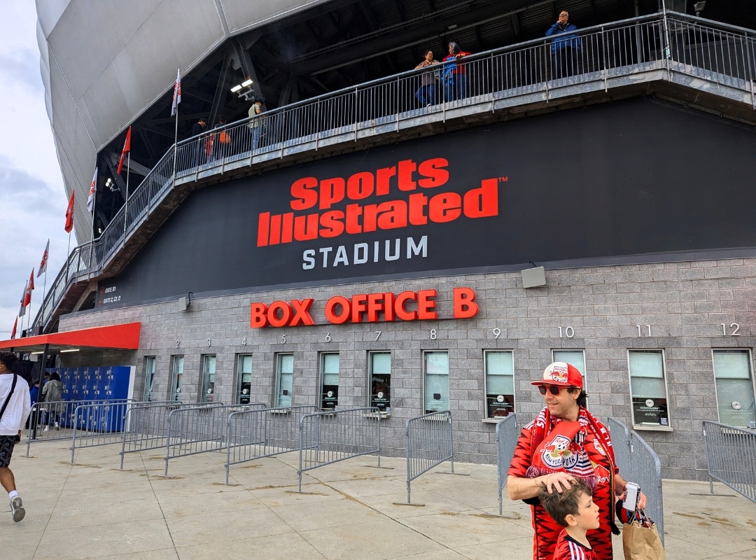 Exterior view of Sports Illustrated Stadium showing Box Office B windows 1 to 10; a man and child in sports attire are in the foreground.