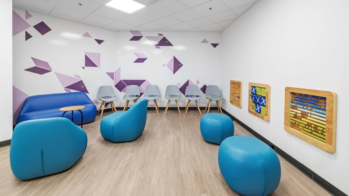 A waiting room with blue chairs and beanbags, a blue couch, a small table, wall-mounted activity boards, and geometric wall decals on white walls.