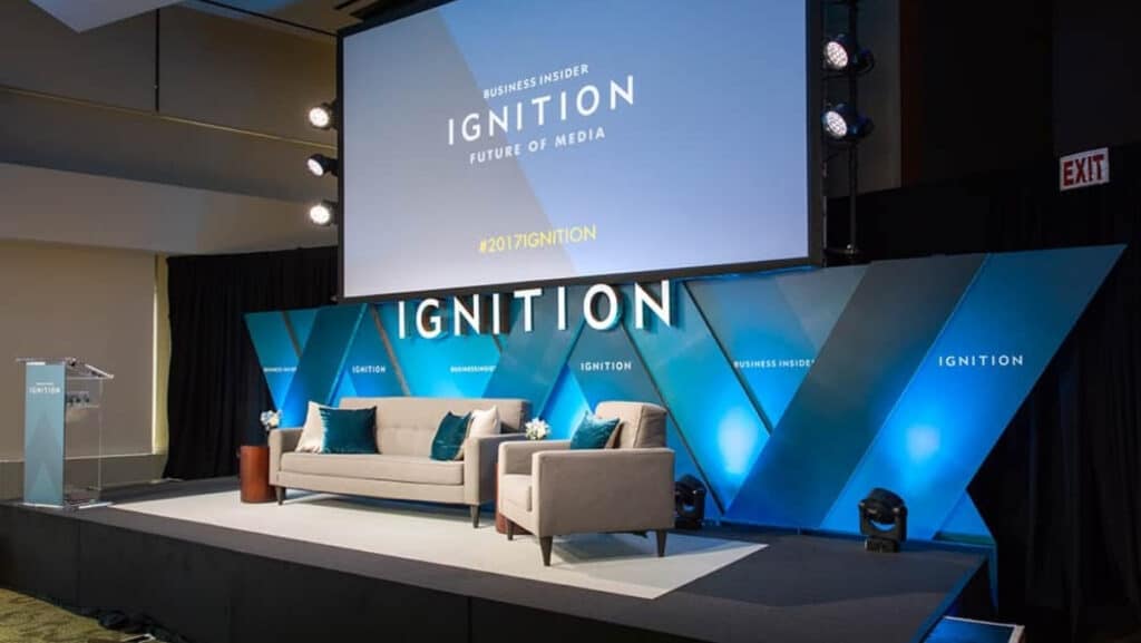 Webstie 50 50 modules (2) A modern stage setup for the Business Insider IGNITION: Future of Media event, featuring a large screen, sofas, chairs, and blue geometric backdrop panels.