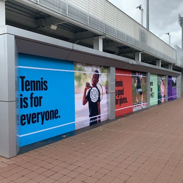 A row of colorful wall panels by a tennis court displays messages promoting tennis inclusivity, including "Tennis is for everyone.