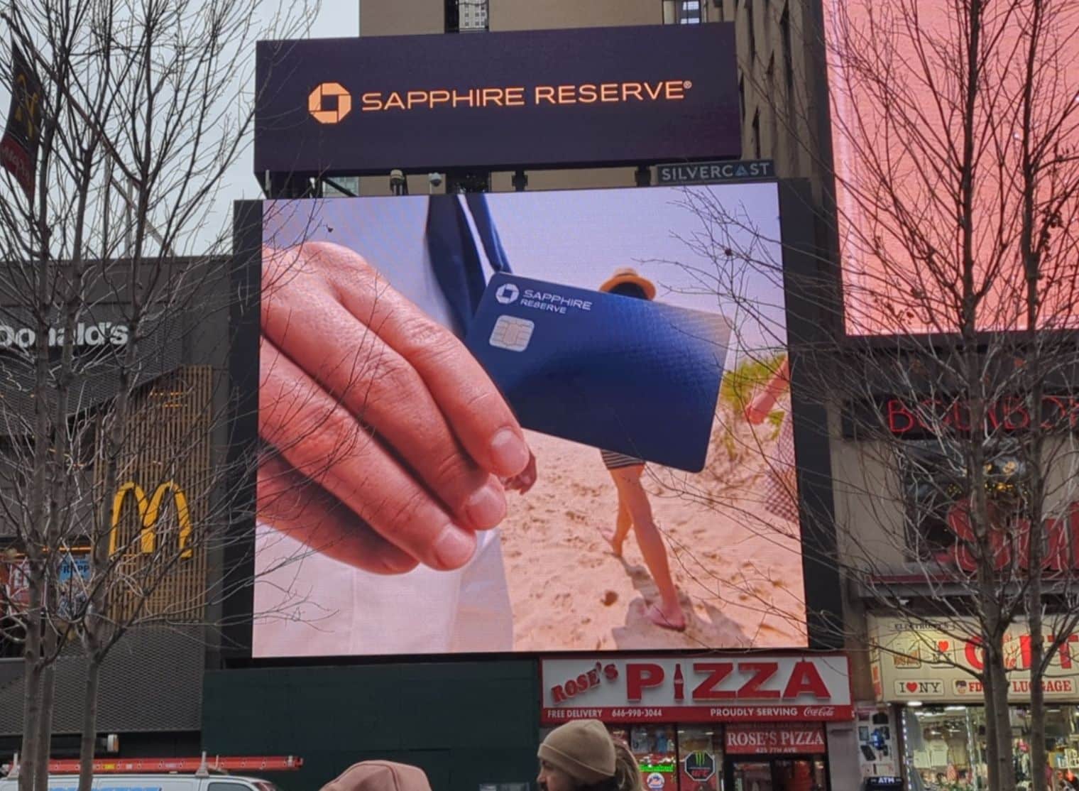 A large digital billboard displays a close-up of a hand holding a Sapphire Reserve credit card with a beach in the background. McDonald's and Rose's Pizza signs are visible below.