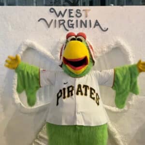 A green mascot in a "Pirates" baseball jersey stands with arms outstretched in front of a "West Virginia" sign on a white wall.