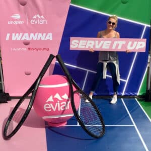 A woman in sunglasses holds a "Serve It Up" sign next to two oversized tennis rackets and a large pink Evian tennis ball at a US Openand Evian promotional display.
