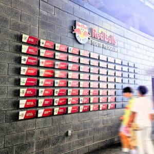 A wall display for Red Bulls Youth Programs with rows of sponsor plaques; two people walk past in the foreground.