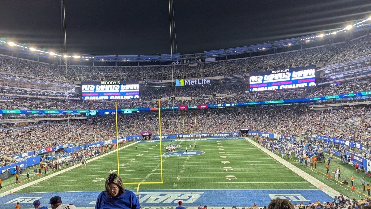 A large crowd fills MetLife Stadium during a night football game, with players on the field and stadium lights illuminating the scene.