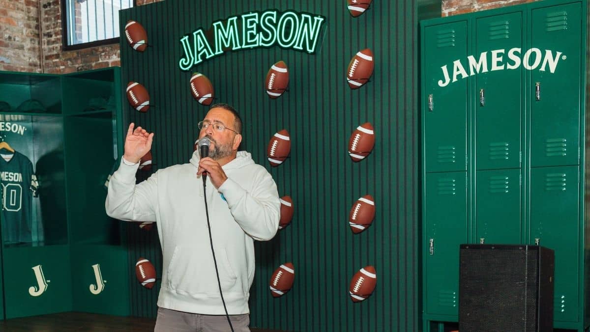 A man speaks into a microphone in front of a green wall decorated with footballs and "Jameson" logos. Lockers and a jersey are visible in the background.