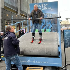 A man in ski gear stands on an indoor ski simulator, holding onto a rail, while another man in a jacket labeled "TEAM" assists him. A West Virginia backdrop is displayed behind them.