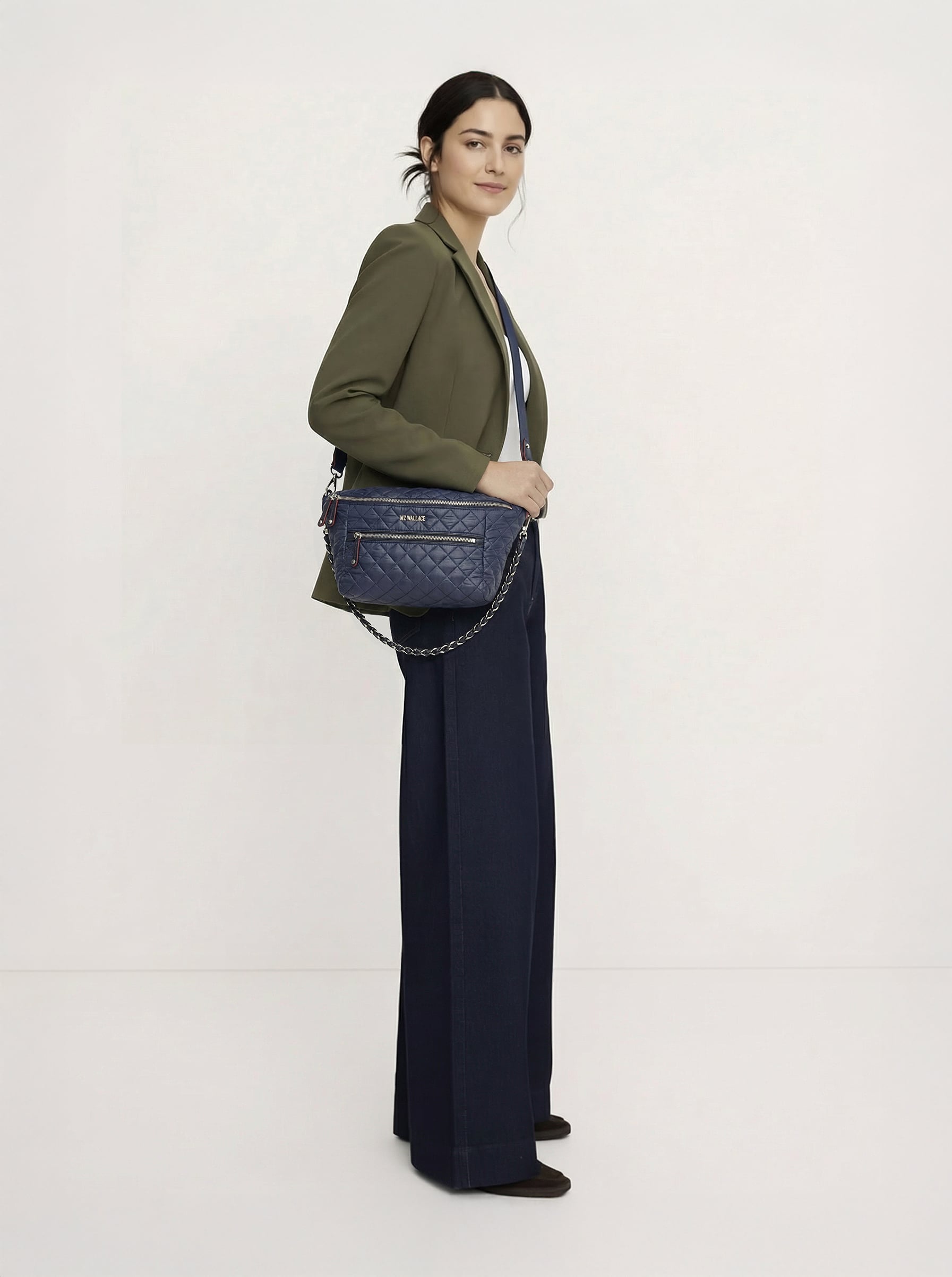 A woman stands against a plain background wearing a green blazer, dark wide-leg pants, brown shoes, and carries a blue quilted shoulder bag.