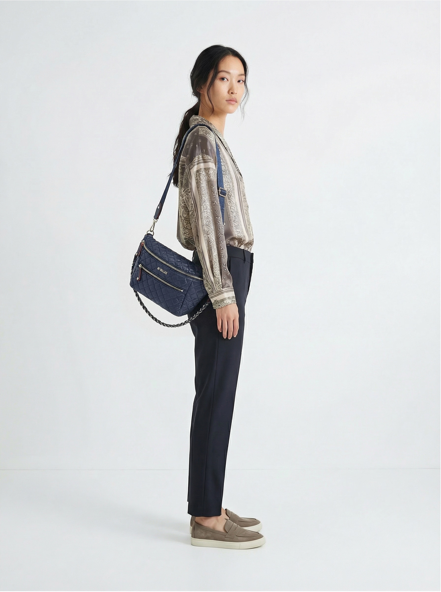 A woman stands in profile against a plain background, wearing a metallic blouse, dark trousers, slip-on shoes, and carrying a blue shoulder bag.