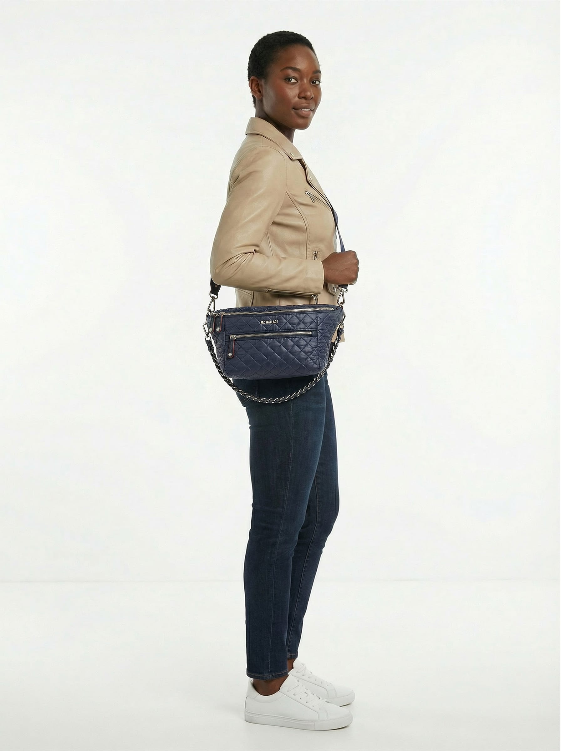 A person stands in profile wearing a beige jacket, dark jeans, white sneakers, and carrying a navy quilted shoulder bag with a chain strap.
