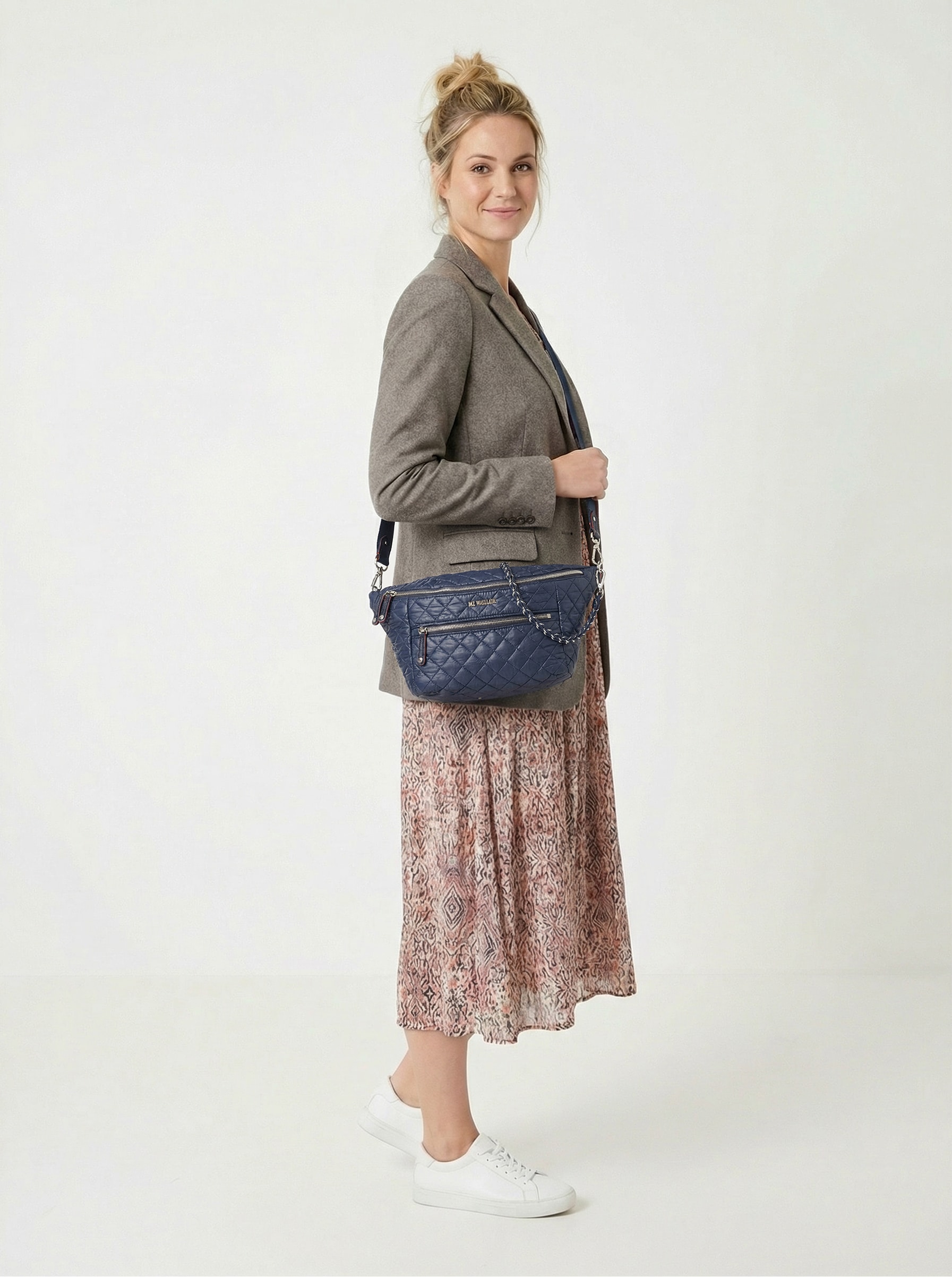 A woman stands sideways wearing a grey blazer, patterned midi skirt, white sneakers, and carrying a navy quilted shoulder bag against a plain background.