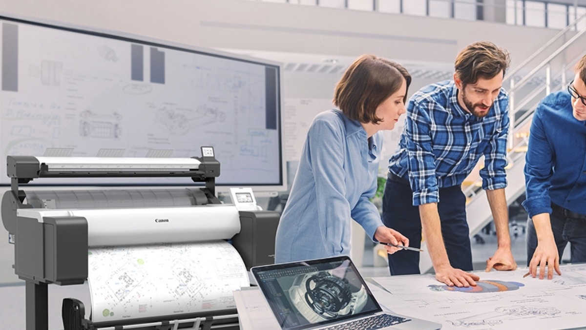 Three people examine technical drawings on a table in an office; a large format Canon printer and a laptop with a mechanical design are in the foreground.