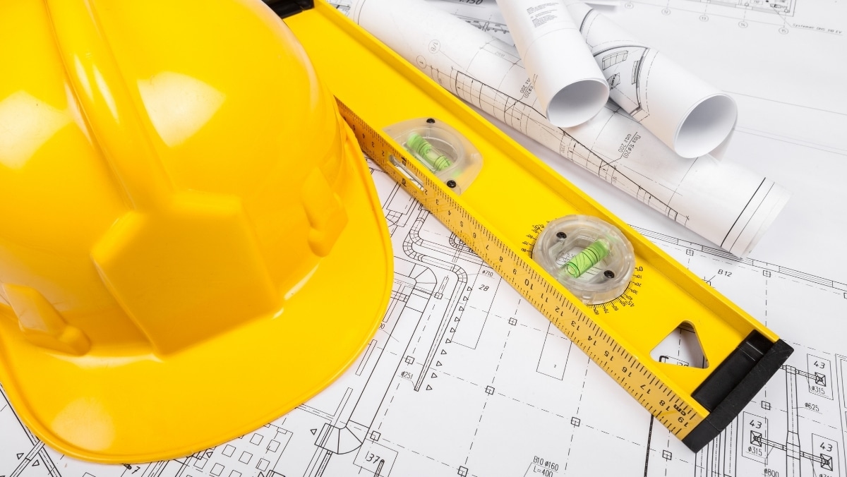 Yellow hard hat, spirit level, and rolled architectural blueprints placed on top of detailed construction plans.