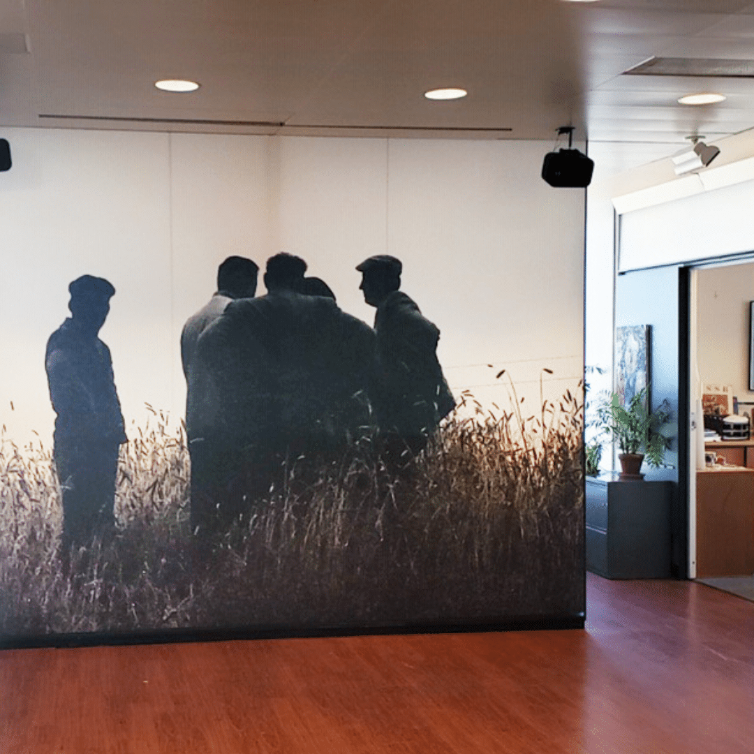 A large black and white mural of five people standing in a field is displayed on an interior wall in a modern room with wood flooring and plants.
