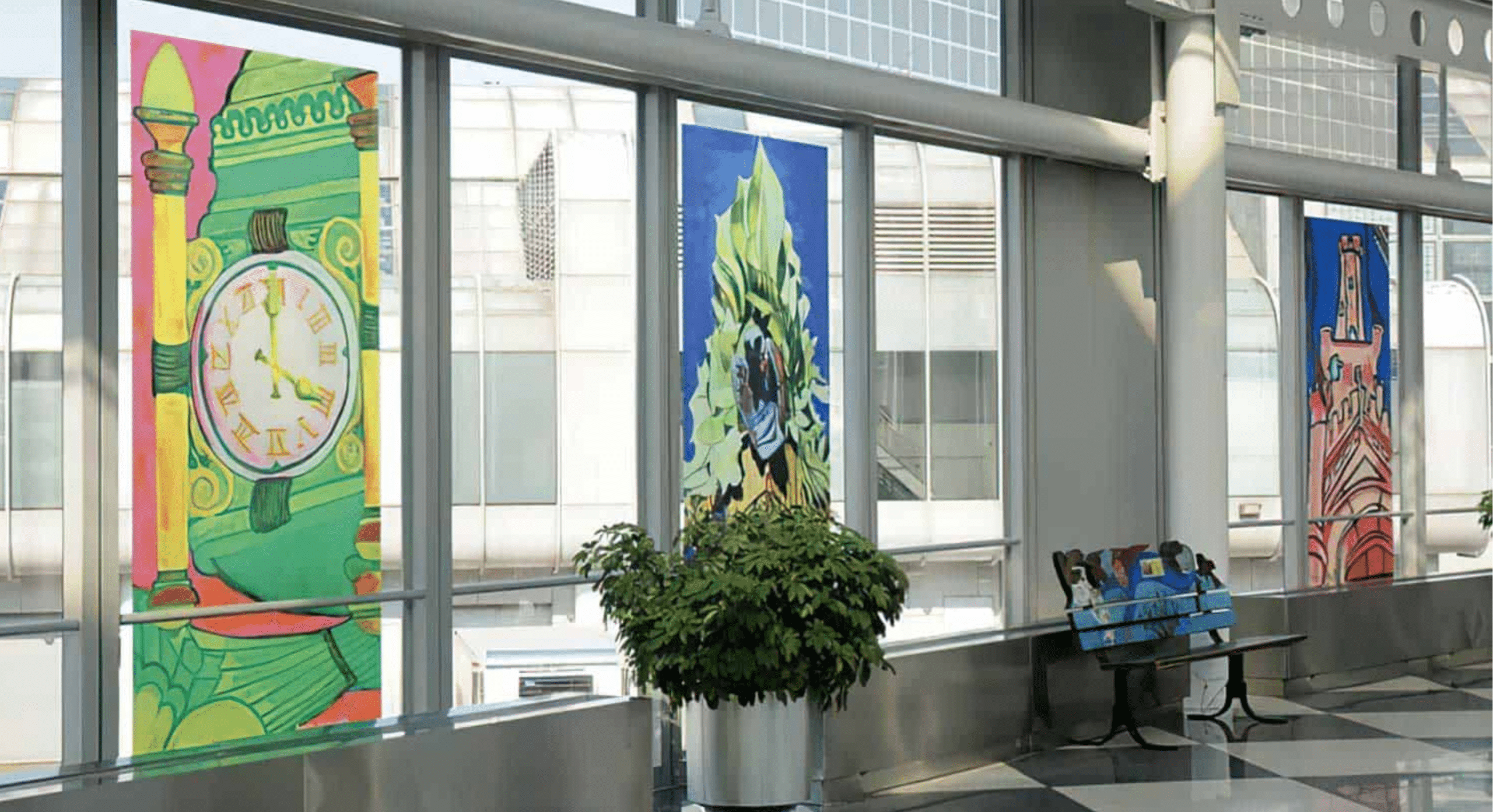 Large, colorful paintings of buildings and a clock are displayed in the windows of a modern indoor space with a potted plant and a bench nearby.