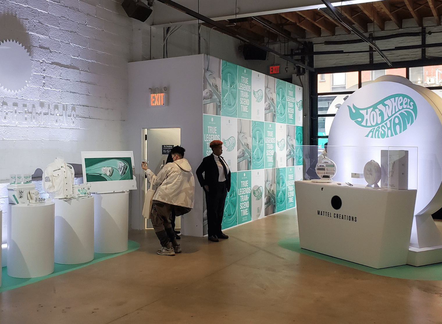 Exhibit space featuring Hot Wheels x Arsham branding, with display cases, informational panels, and two people interacting with the displays.