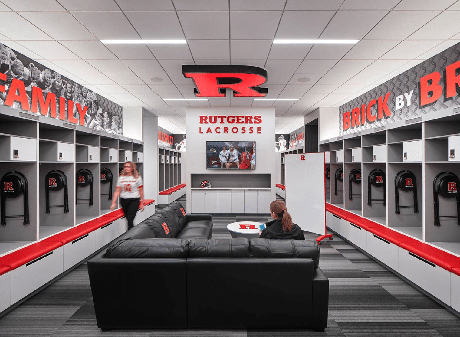 Rutgers lacrosse locker room with black lockers, red accents, a central lounge area, and large Rutgers “R” logos on the walls and ceiling. Two people are present.