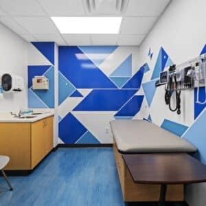 A modern medical exam room with blue geometric wall design, exam table, medical equipment on the wall, and a sink with cabinets.