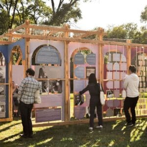 Four people read informational displays mounted on a wooden outdoor exhibit structure in a grassy, tree-lined park under daylight.