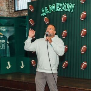 A man in a light hoodie speaks into a microphone in front of a Jameson display decorated with footballs.