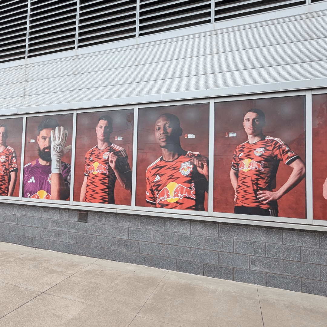 A row of large posters displays five soccer players in red and black New York Red Bulls jerseys on an exterior wall.