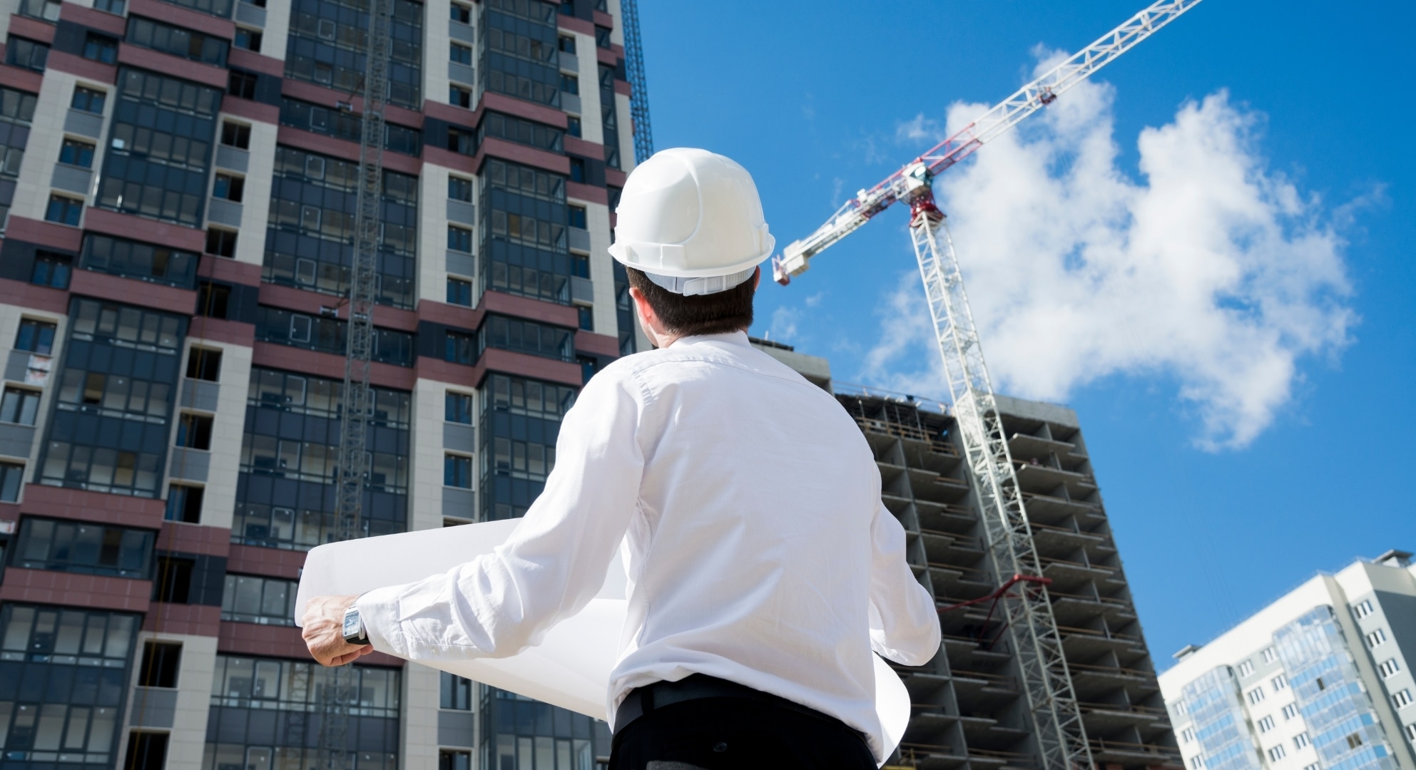 A person in a white hard hat holds blueprints while looking at a construction site with a tall building and a crane against a blue sky.
