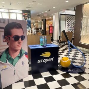 Display for the US Open featuring a large tennis racket and ball, a poster of a man in sunglasses, and promotional materials on a checkered floor inside a retail store.
