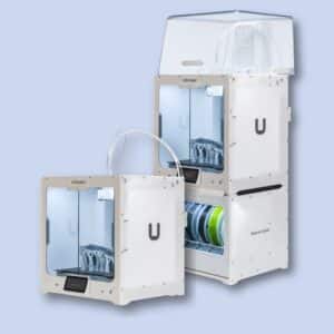 Two white Ultimaker 3D printers are shown side by side, each with transparent doors and spools of filament visible inside or below the machines.