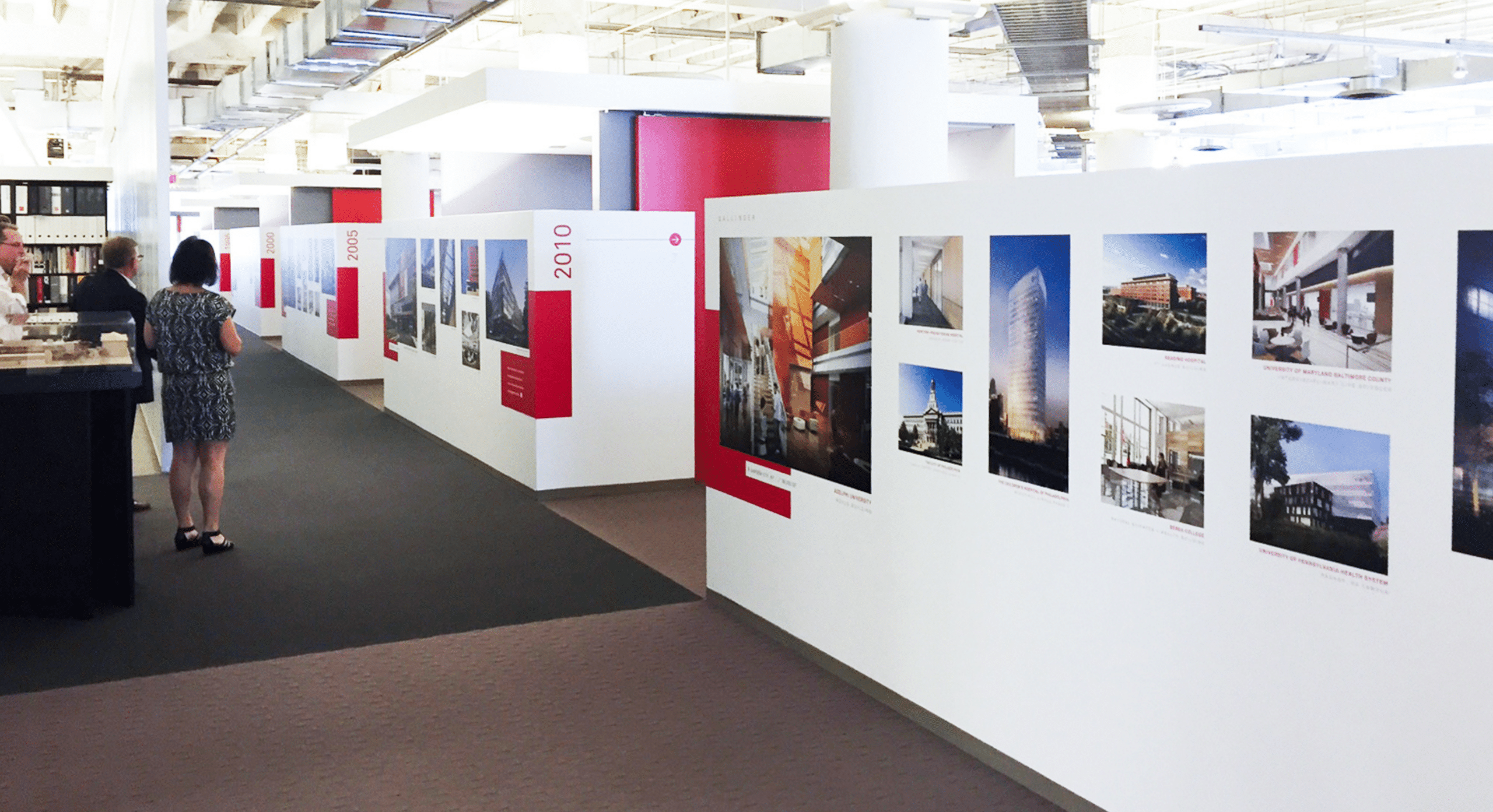 Two people view a hallway display featuring large panels with architectural photos and captions, arranged chronologically along a brightly lit office space.