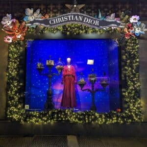 A Christian Dior store window display features a mannequin in a pink dress, birdcages, and a blue starry background, framed by festive greenery and lights.